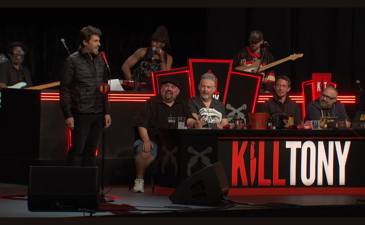 Adam Ray onstage as Tony Hinchcliffe at the Intuit Dome, with the real Tony and the panel at the desk, Kill Tony episode 780.