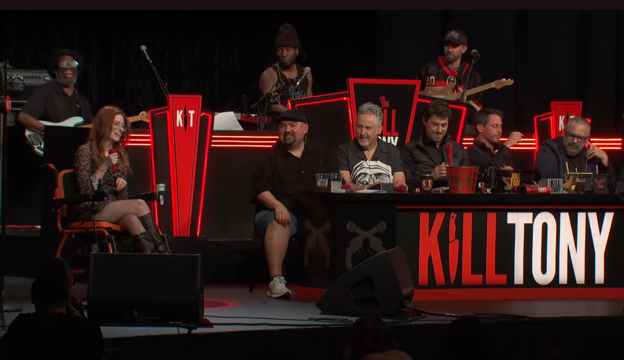 Fiona Collie onstage in a wheelchair with the Kill Tony panel and band behind her, Intuit Dome, episode 780.