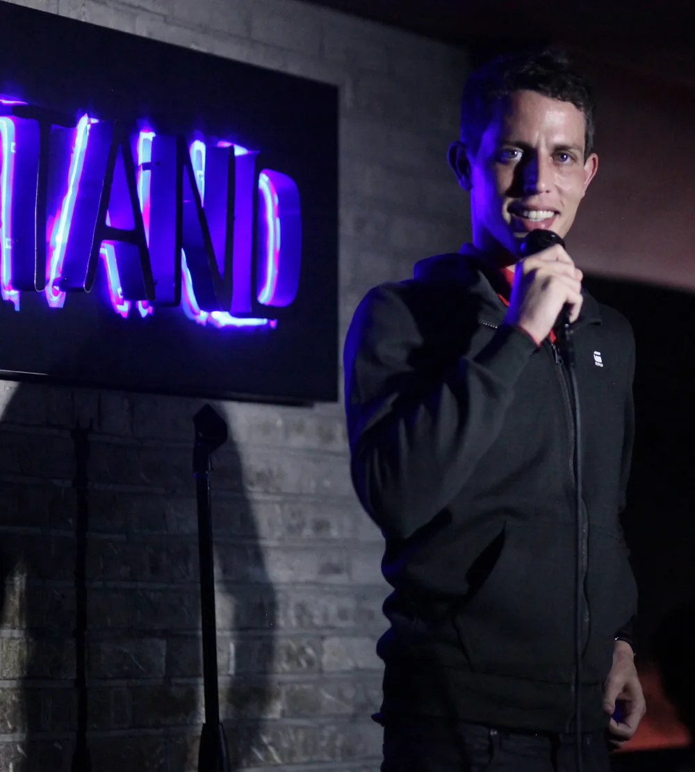 Tony Hinchcliffe performing stand-up with a microphone, neon Stand sign behind him, August 2017.